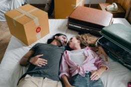 A couple lying on the bed with a cardboard box and suitcases